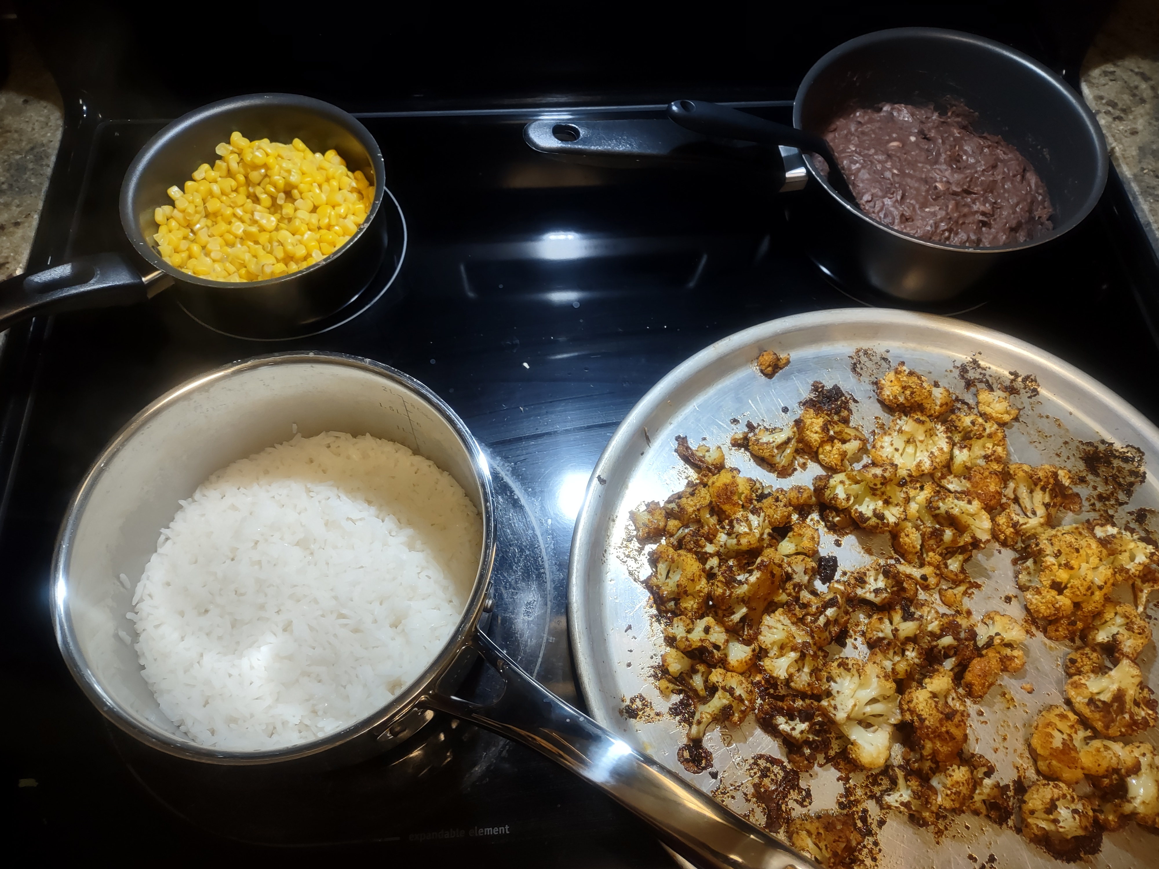 photo of three saucepans and a pizza pan containing corn, refried black beans, rice, and roasted taco seasoned cauliflower